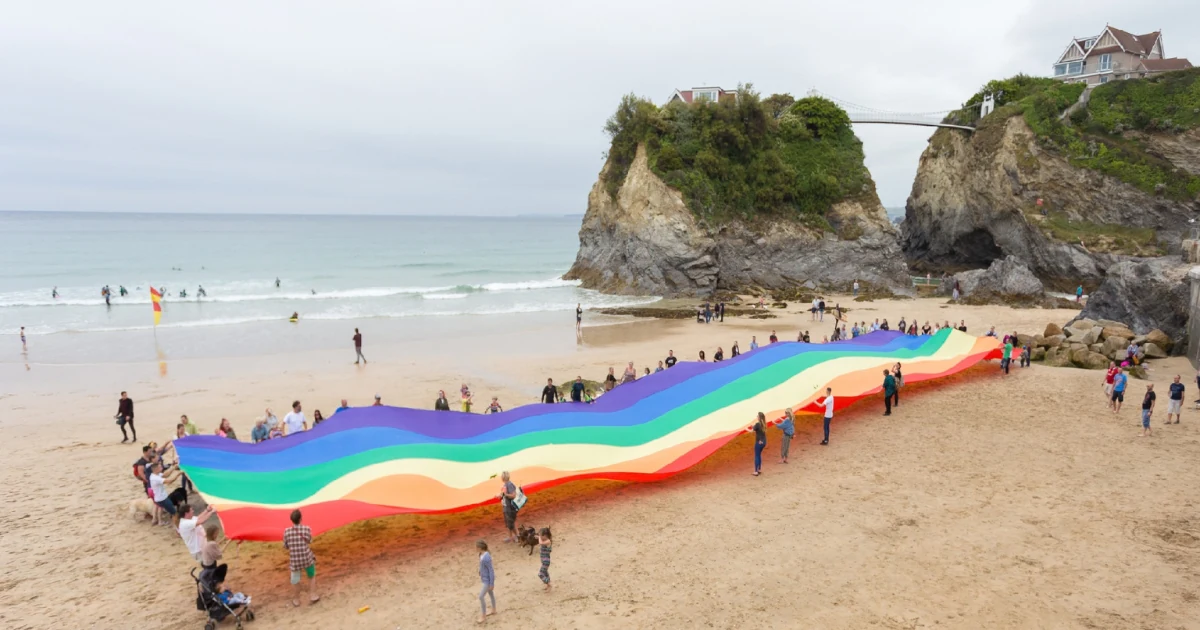 People holding a long rainbow Pride flag across a Cornish beach beside the sea and cliffs